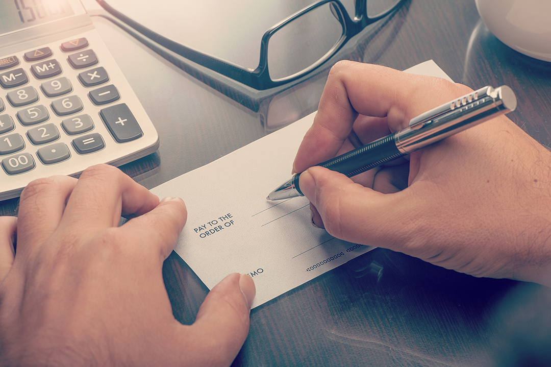 Close up of hands writing out a check with a calculator and pair of glasses in the background