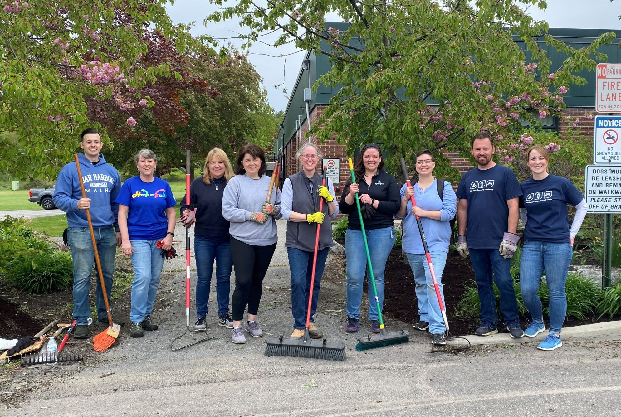 Photo of a team of volunteers posing with rakes and brooms in front of a building and lawn.