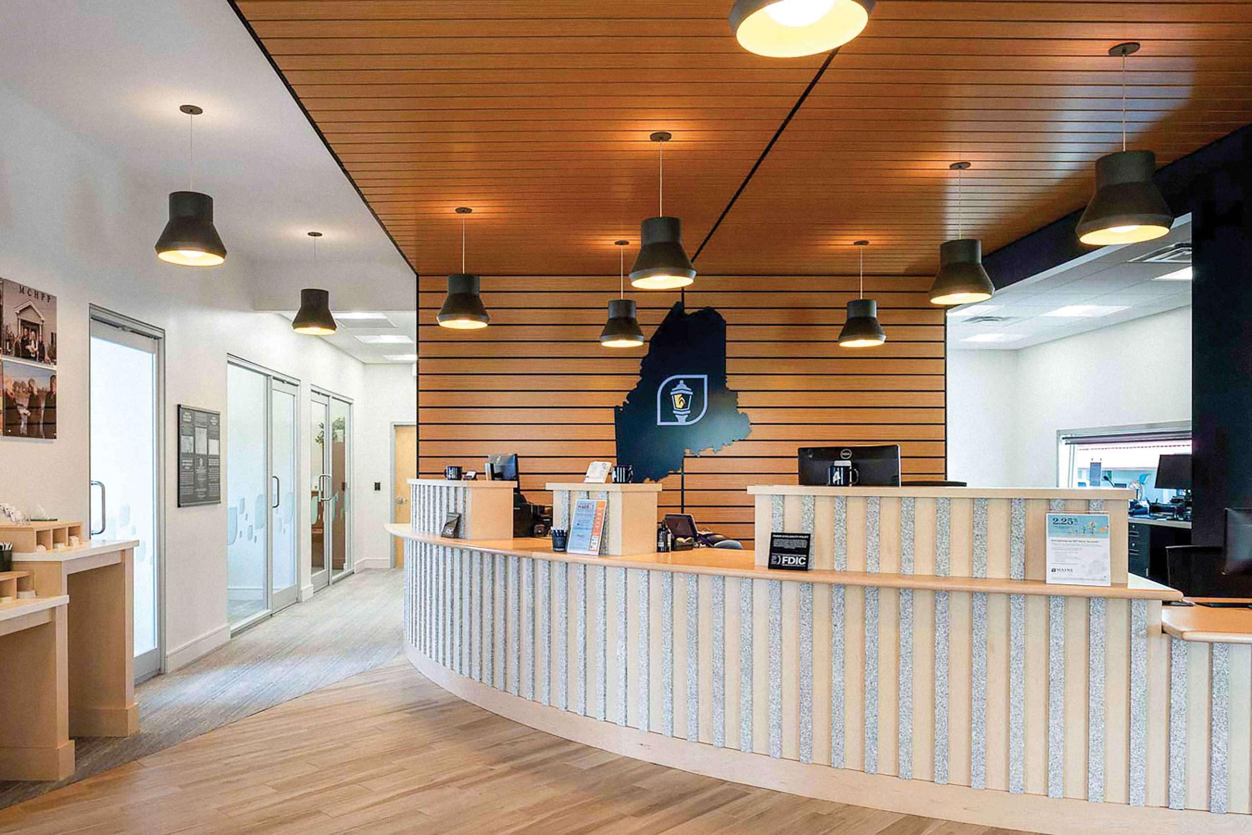 A modern, well-lit Maine Community Bank branch reception area with a curved desk, wood accents, hanging lights, and a wall featuring a map outline and emblem behind the counter.