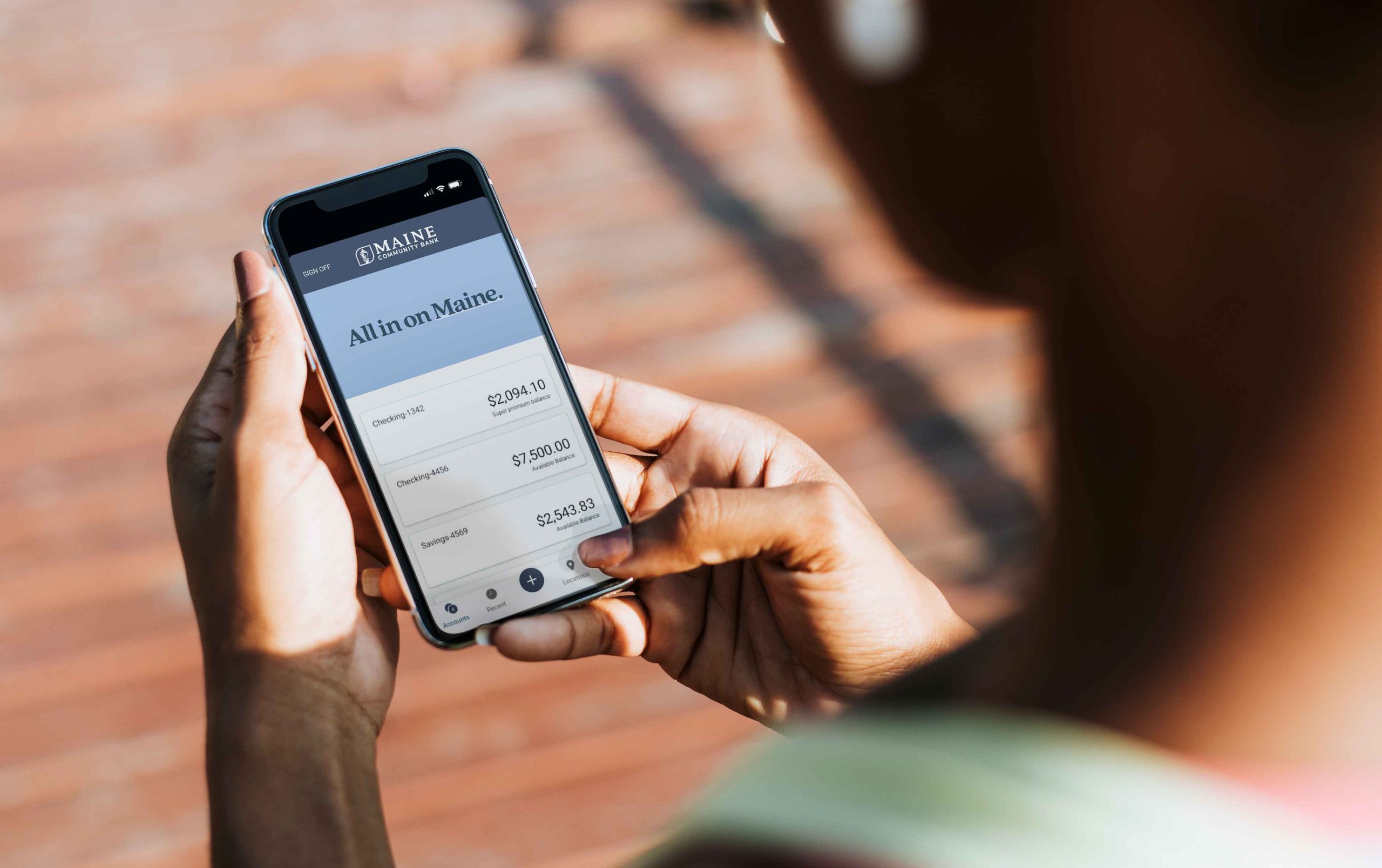 A person is holding a smartphone displaying a Maine Community Bank's mobile app. The screen shows the 'Maine Community Bank' logo and the text 'All in on Maine.' Below, three account balances are listed: 'Checking 1' with $2,094.10, 'Checking 2' with $7,500.00, and 'Savings 1' with $2,543.85. The person's thumb is touching the screen near the bottom right corner.