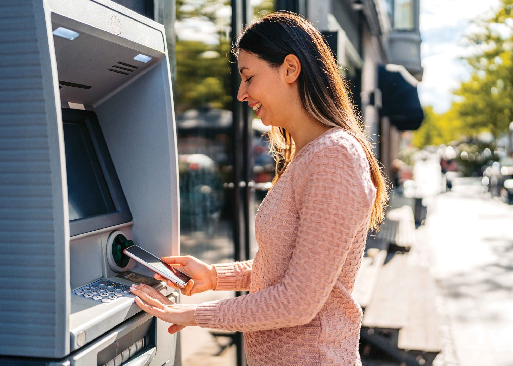 A person in a pink sweater is using an ATM machine on a sunny day. The person is holding a bank card and appears to be inserting it into the machine. The background shows an outdoor urban setting with trees and buildings.