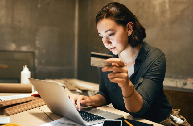A woman sits at a table using a laptop while holding a credit card, appearing to make an online purchase.